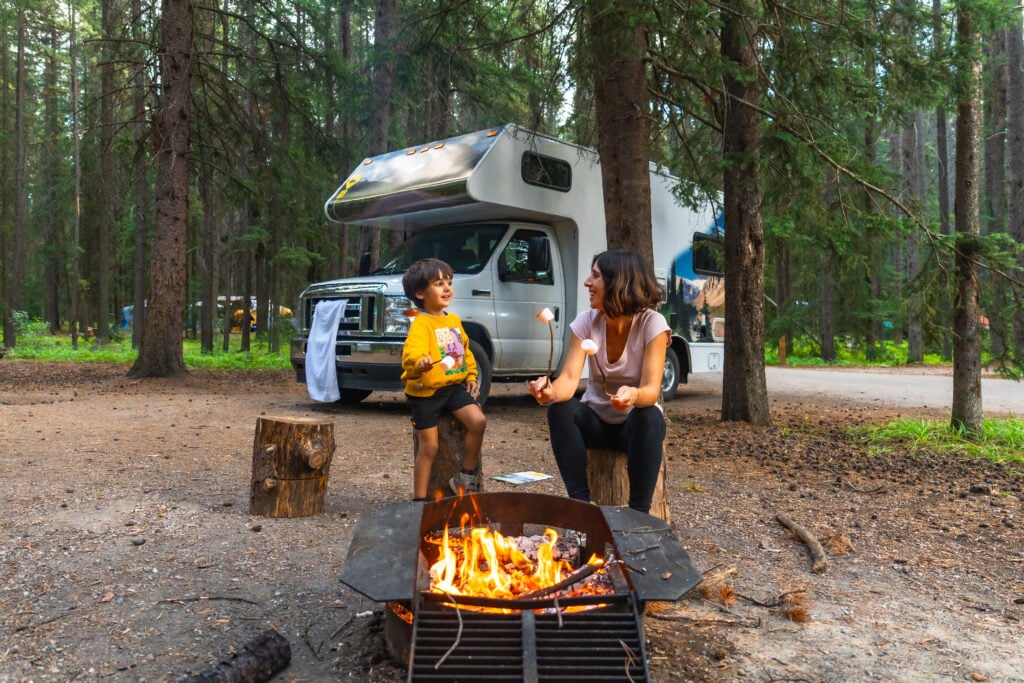 Family camping with marshmallows by campfire and RV.