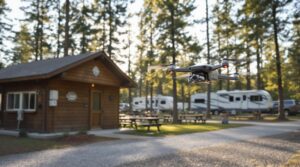A drone with a camera hovers beside a generic wooden campground utility building, surrounded by picnic tables, gravel path, and blurred trees in the background on a sunny afternoon.